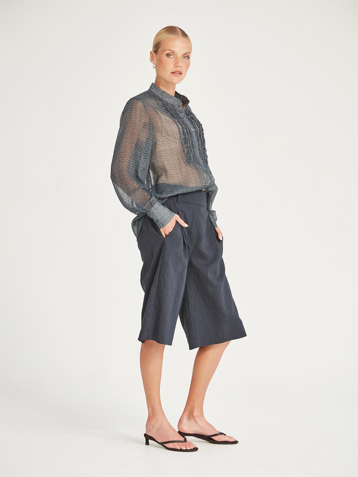 Side view of the Caroline Sills Dauphine Blouse in navy print, showing a Mandarin collar neckline, ruffle trim detail on the bust and sheer long sleeves. Paired with navy shorts.