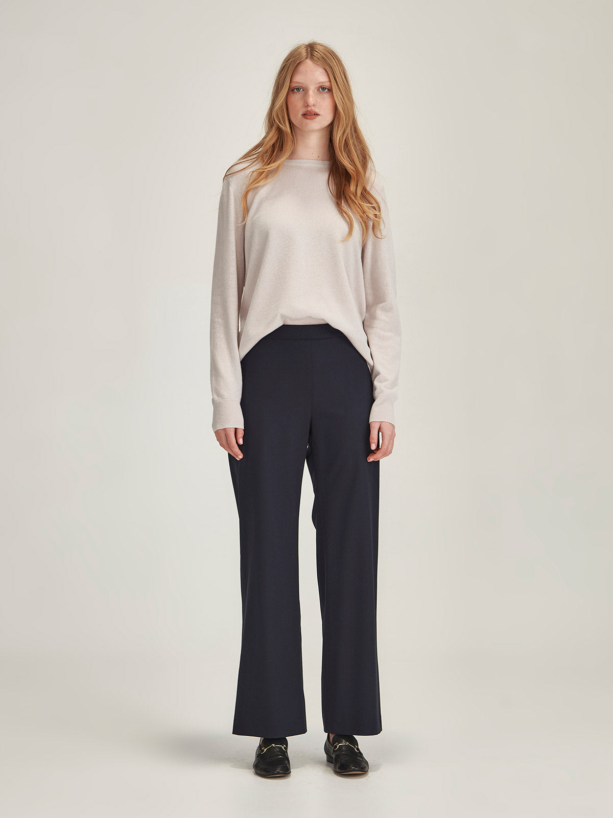 Woman wearing Caroline Sills Eleanor crew neck Cashmere Sweater in Vapour beige and Claude Pant in Navy against grey backdrop
