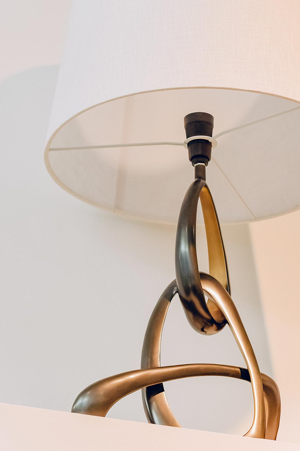 Contemporary bronze coloured metal lamp with a curved design and white fabric shade, displayed on a shelf in-store at Sills + Co.