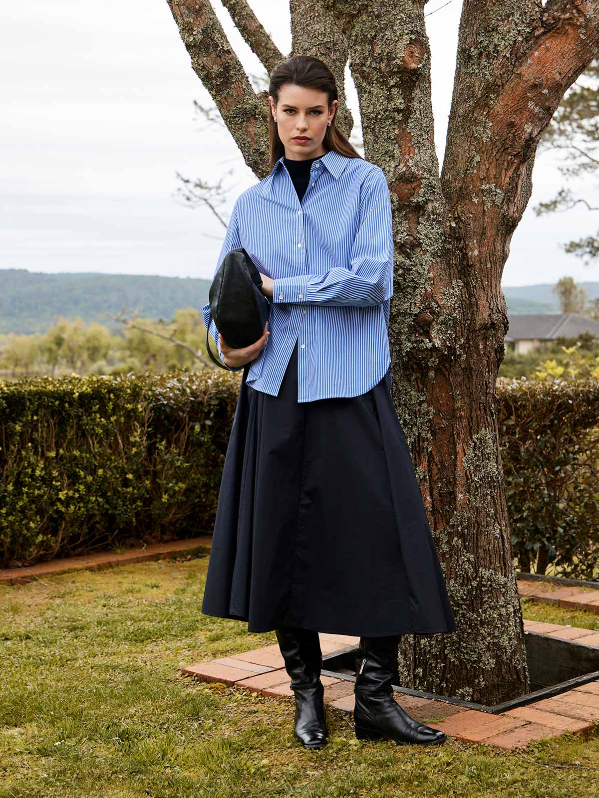 women outdoors by tree wearing Sills navy blue Princeton Striped Shirt, black Zaiya Godet midi Skirt, and Juju & Co Sling Bag