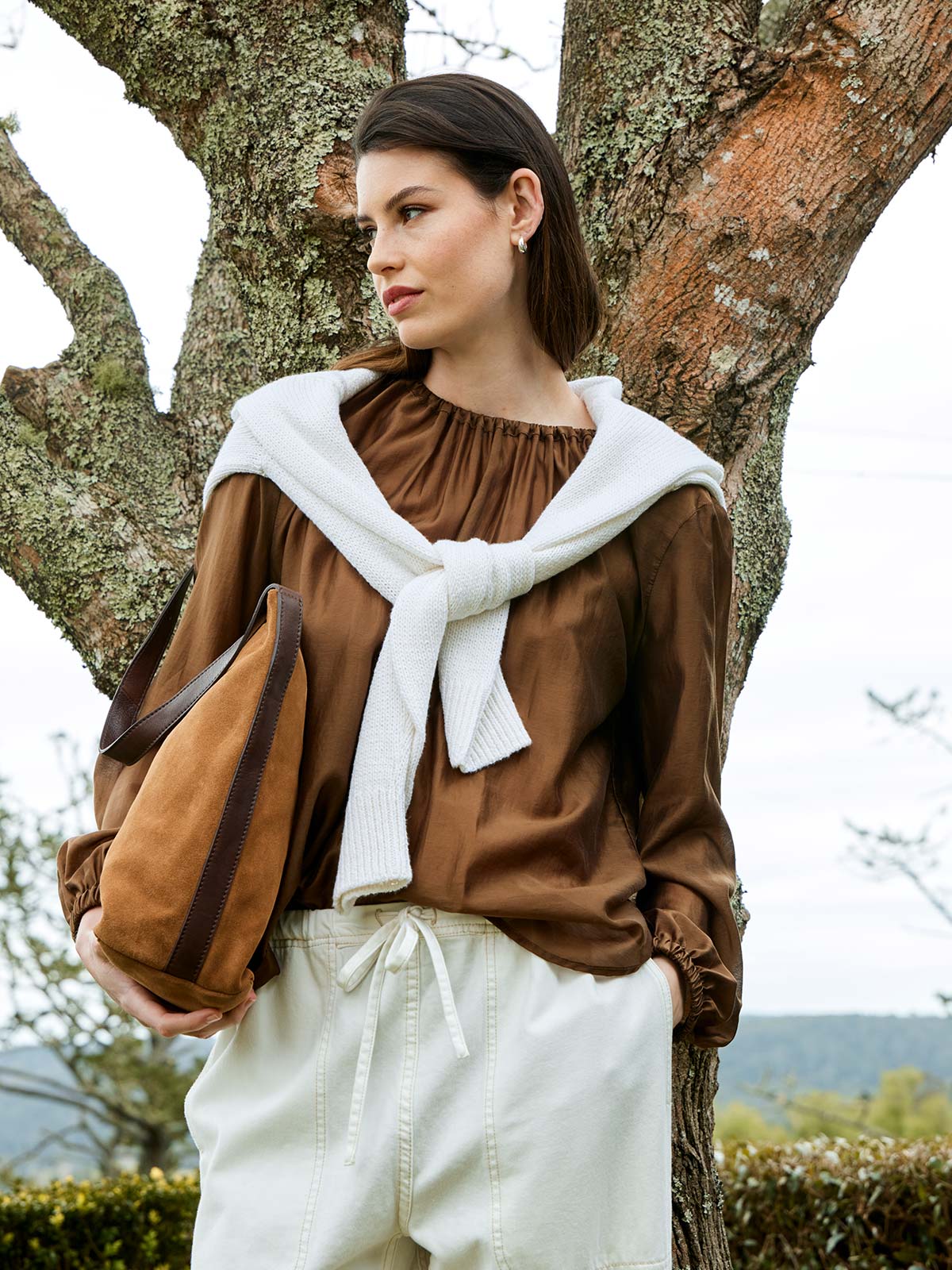 woman outdoors wearing Sills Cinnamon Anna Top bronze chiffon blouse with sweater around shoulders and Juju & Co suede bag