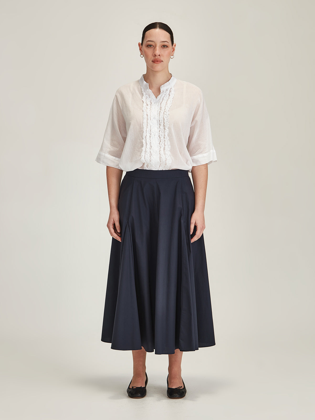Model wearing Sills Sasha Ruffle blouse in white organic cotton and Zaiya navy midi skirt in front of grey background