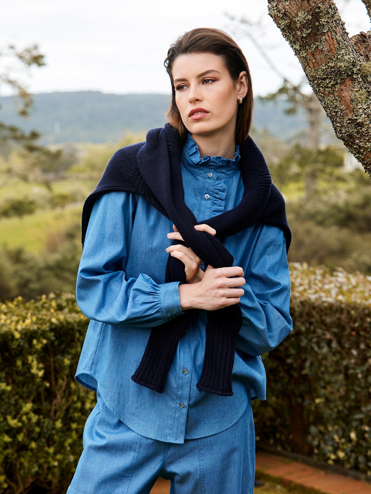 A woman outdoors wearing the Sills Mira Ruffle Blouse in chambray light blue denim with navy sweater around shoulders