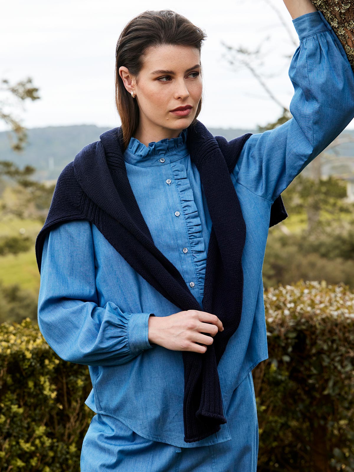 A woman outdoors wearing the Sills Mira Ruffle Blouse in chambray light blue denim with navy sweater around shoulders