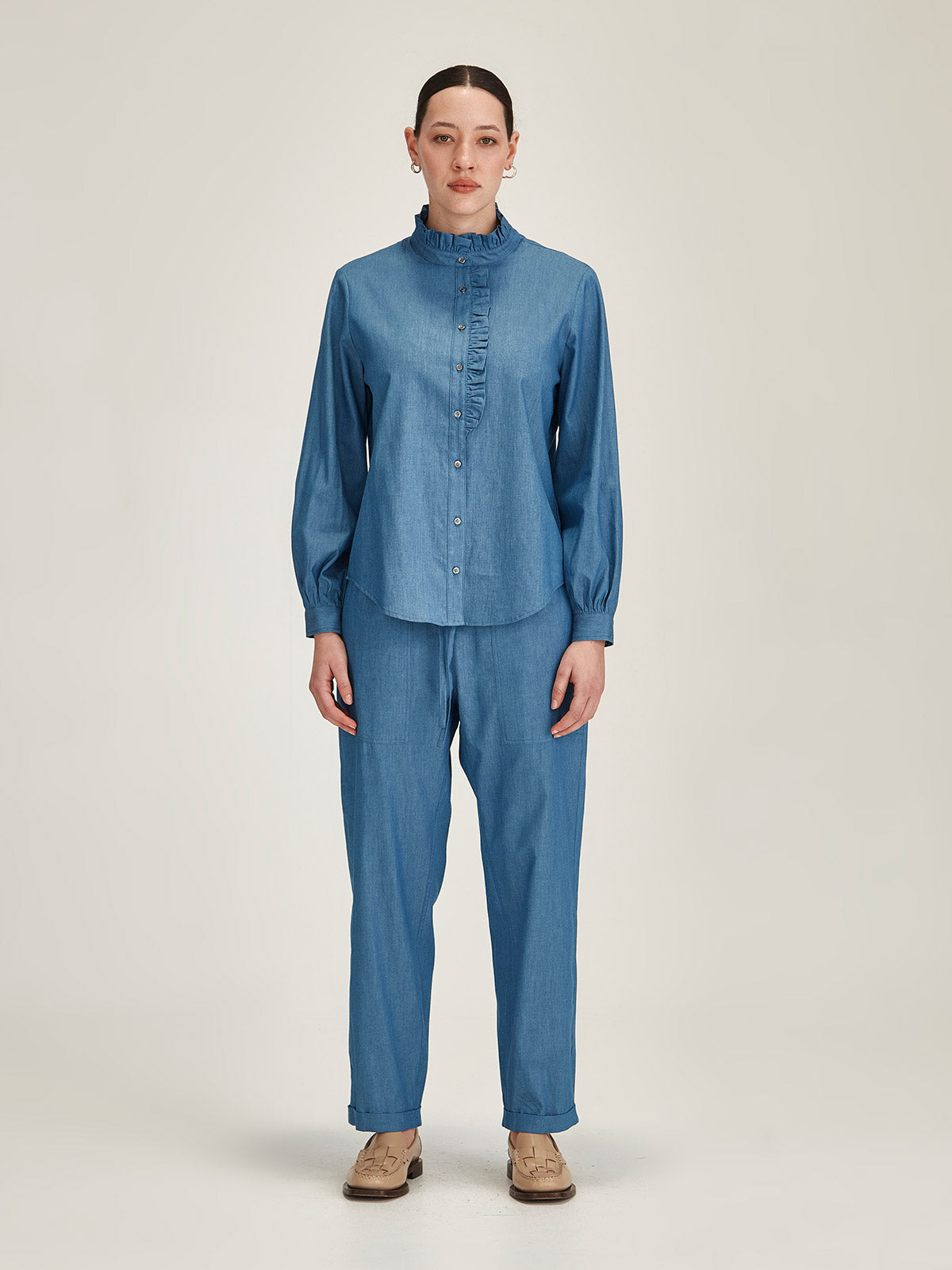 Woman wearing matching chambray Sills Mira Ruffle Blouse and Serena Chambray Pant in light indigo in front of grey background