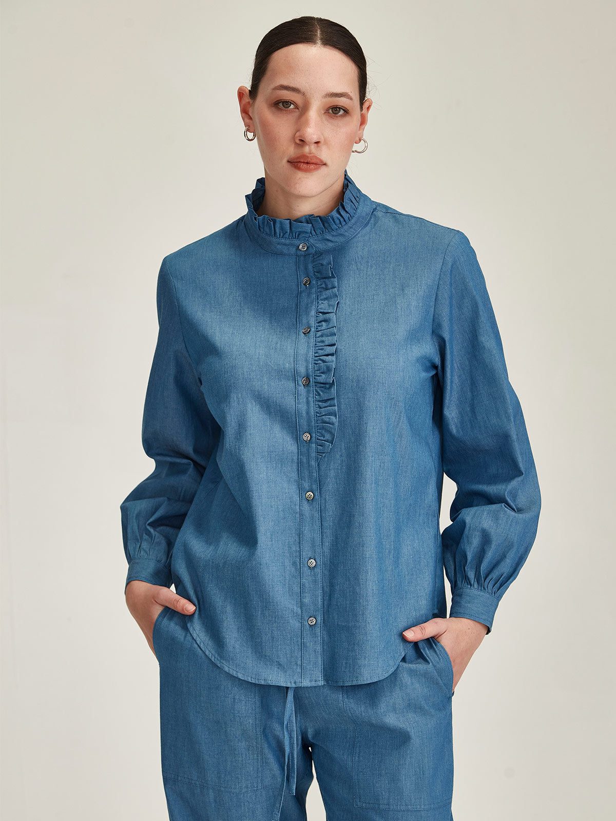 Close up of woman wearing Sills Mira Ruffle Blouse in light indigo chambray with ruffle collar and lapel on grey background