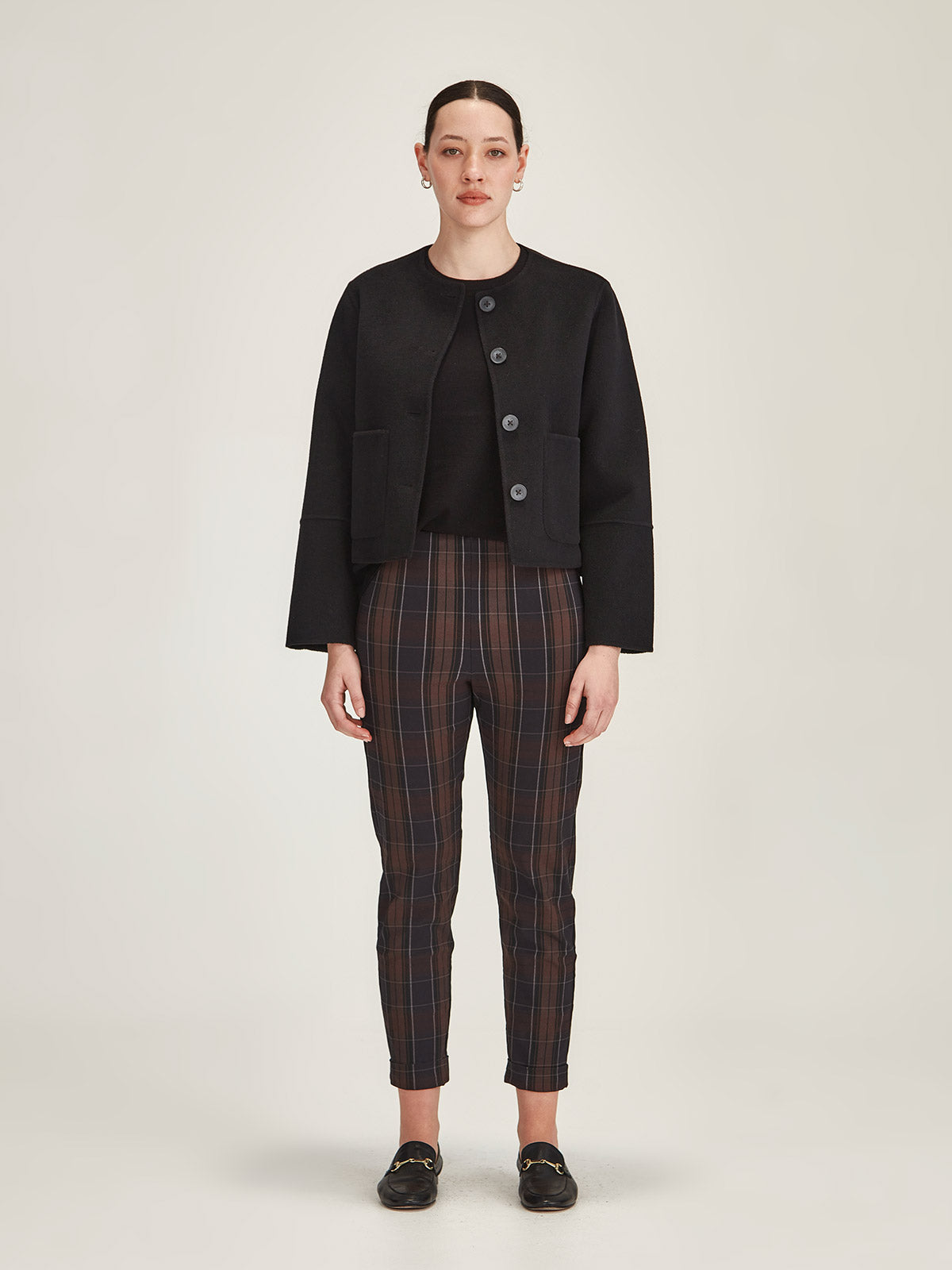 Model wearing Sills Hepburn pants in chocolate plaid check with Emiko wool short coat in black in front of grey background