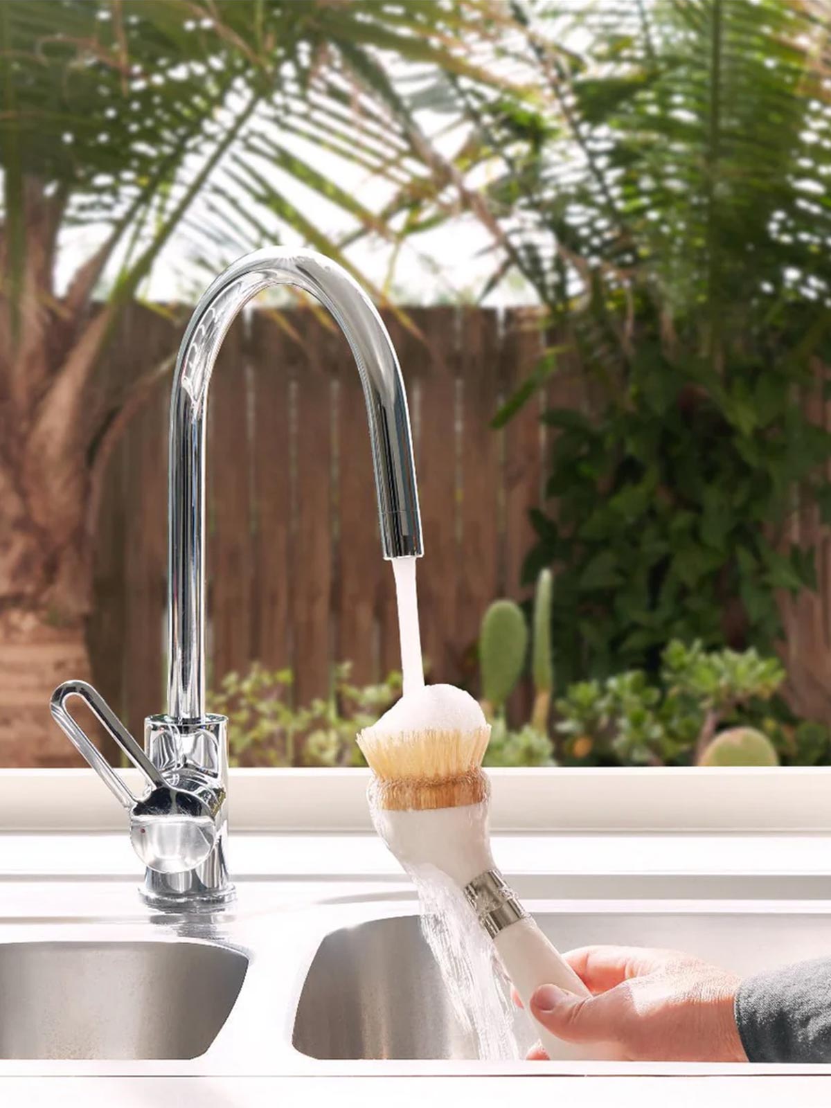 The ONE BRUSH by Yeseco - White eco-friendly dish brush in use, showing compostable sisal fibre bristles being rinsed under a silver kitchen faucet, sustainable kitchen tool with magnetic stand and built-in soap dispenser.