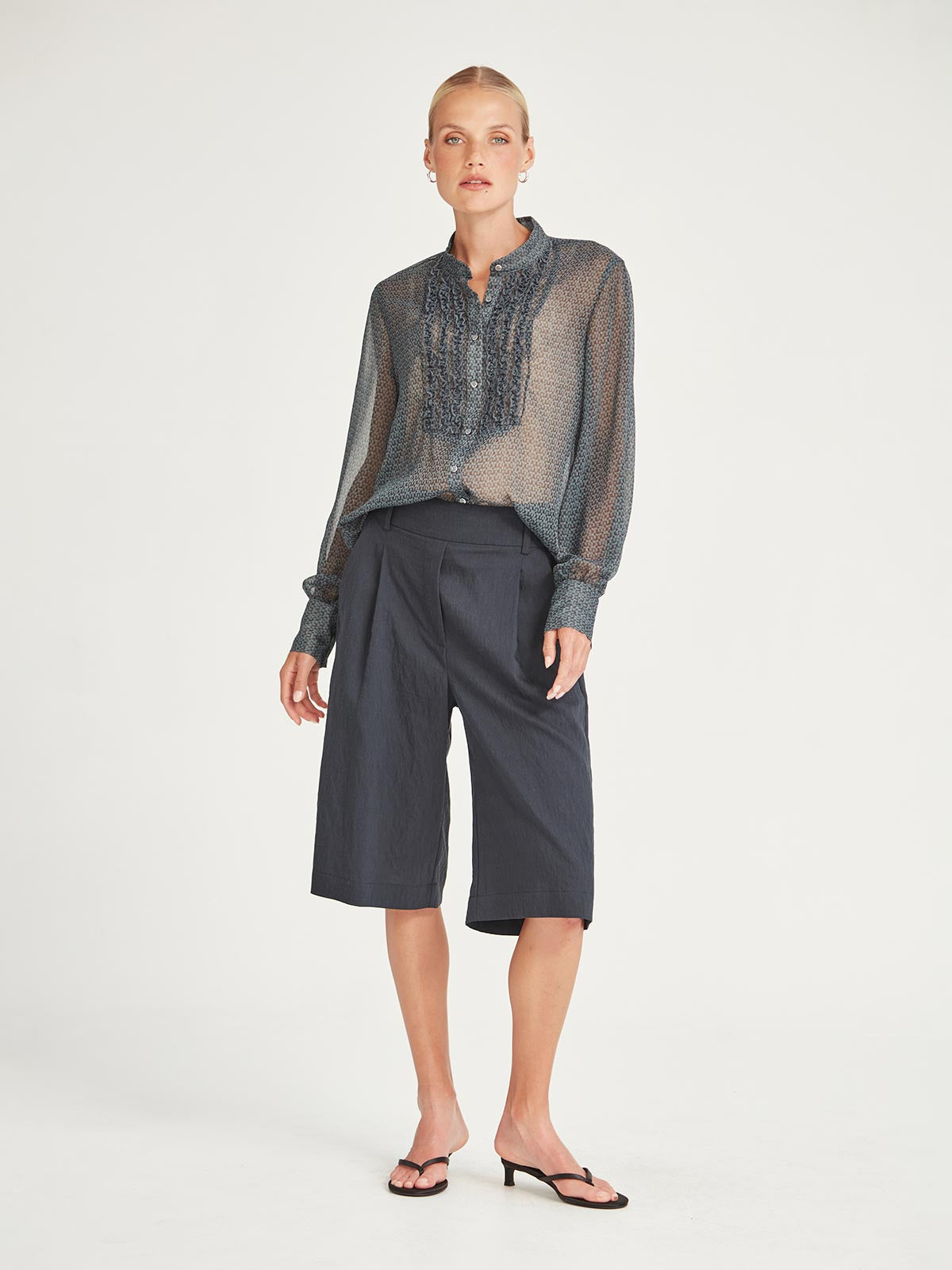 Front view of the Caroline Sills Dauphine Blouse in navy print, featuring a Mandarin collar neckline, ruffle trim detail on the bust and sheer long sleeves. Styled with navy shorts.