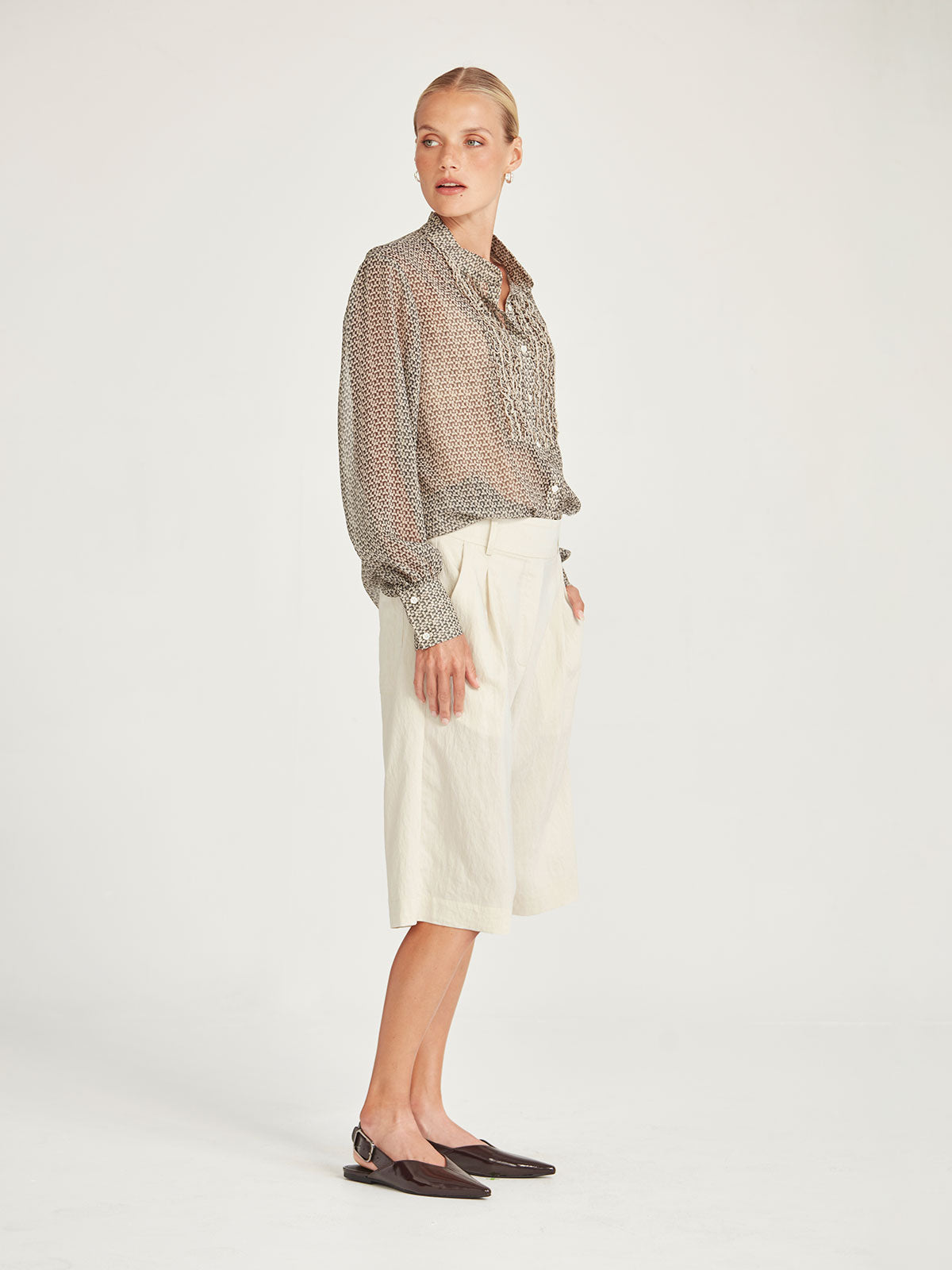 Side view of the Caroline Sills Dauphine Blouse in sand print, showing a Mandarin collar neckline, ruffle trim detail on the bust and sheer long sleeves. Paired with chalk coloured tonal shorts.
