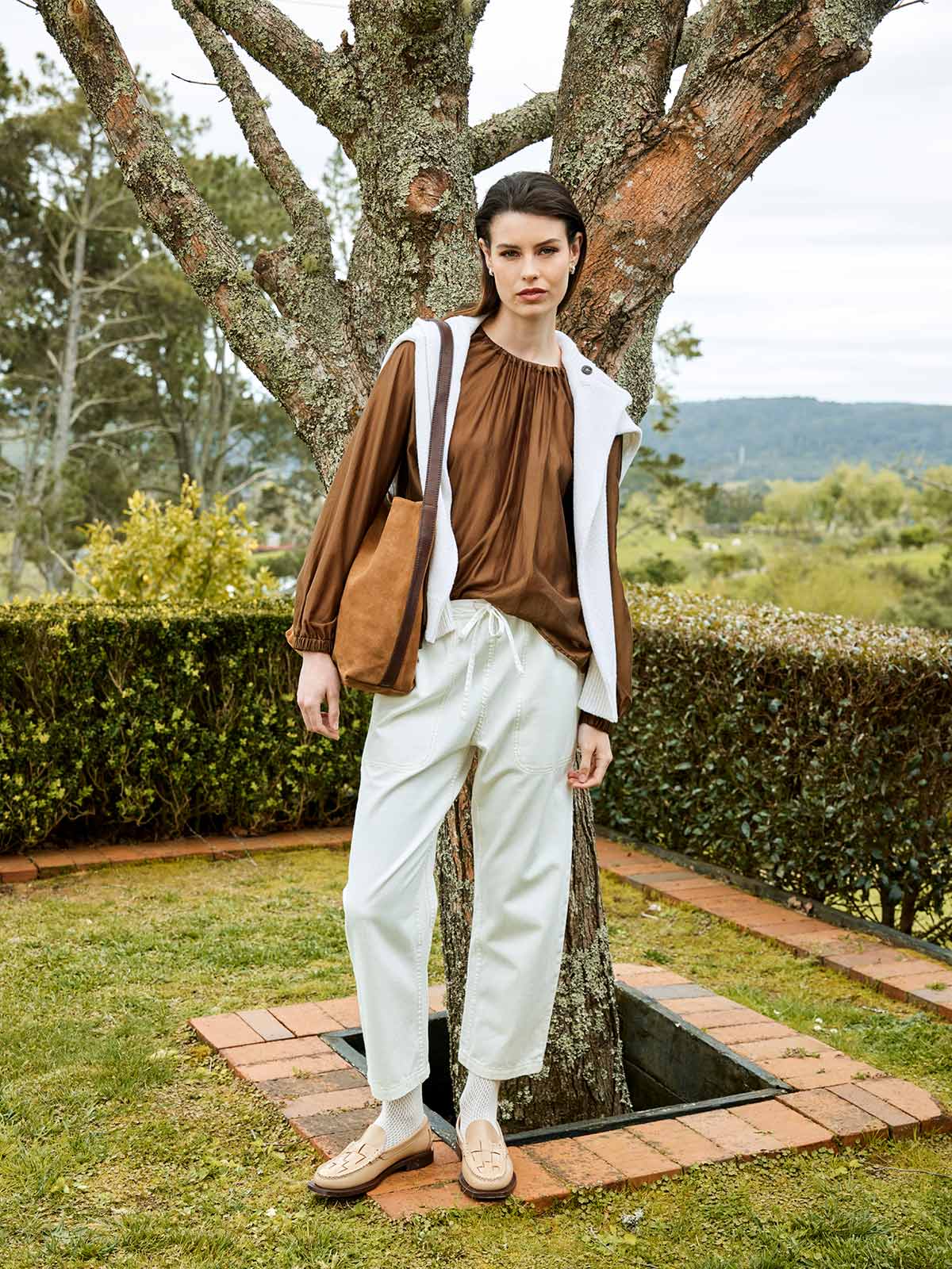 woman outdoors wearing Sills white Ginza Drawstring ankle length Jean with bronze Anna blouse and a knitted sweater over shoulders