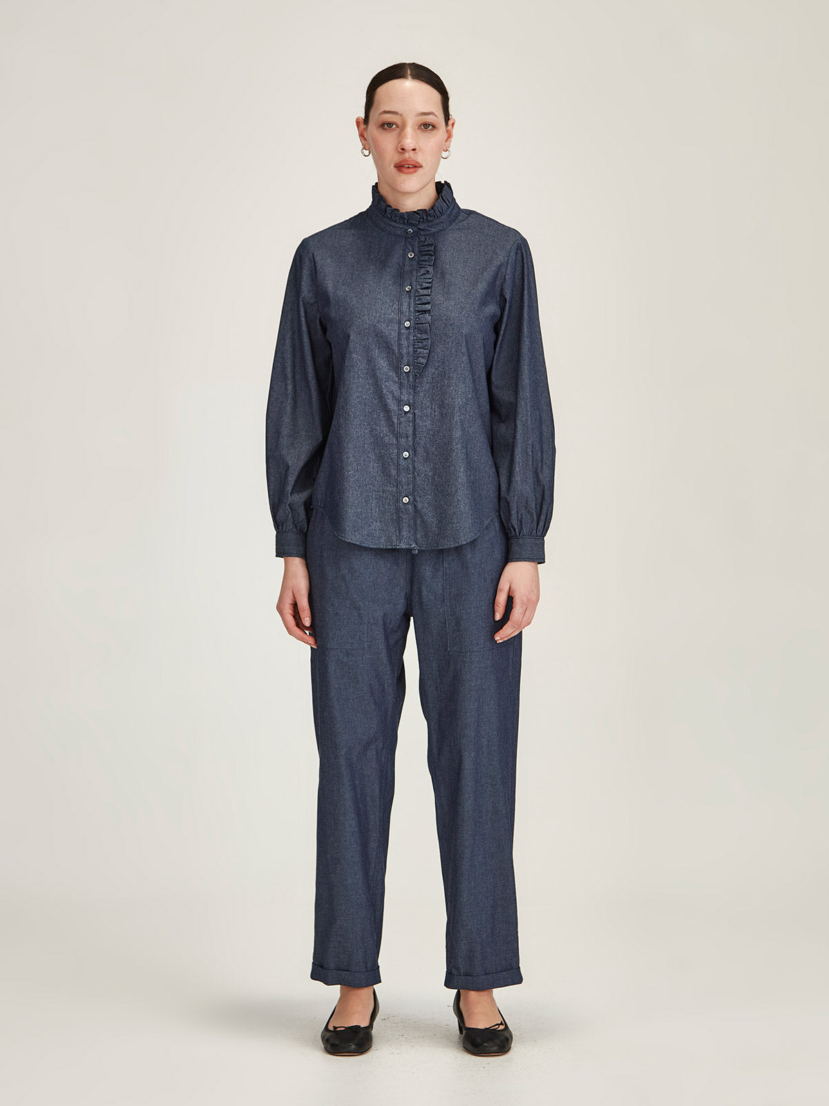 Woman wearing matching chambray Sills Mira Ruffle Blouse and Serena Chambray Pant in dark indigo in front of grey background