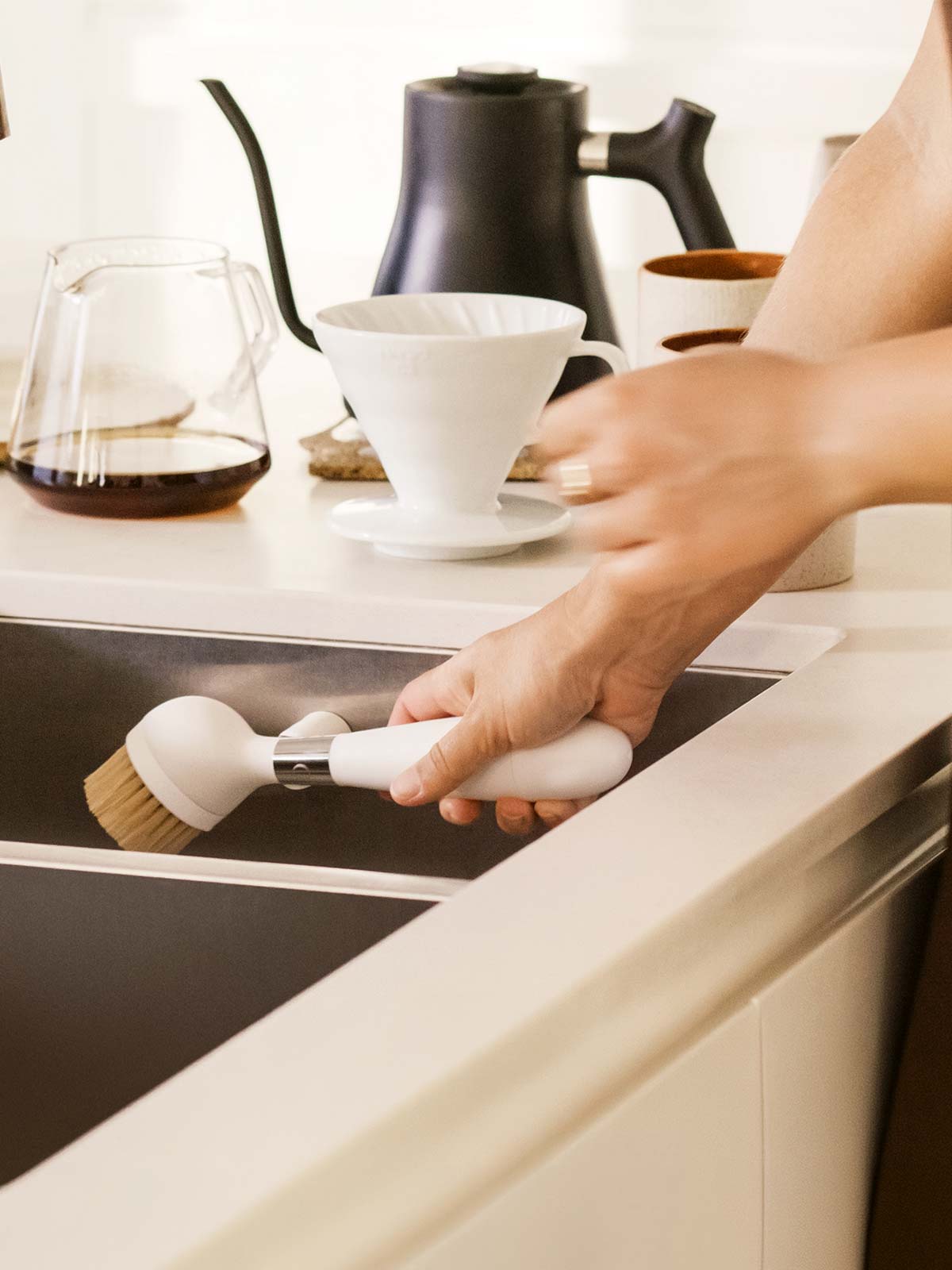 The One Brush V2 Magnetic by Yeseco - Hand holding the white brush beside a dark undermount sink in a modern kitchen with coffee equipment in the background.