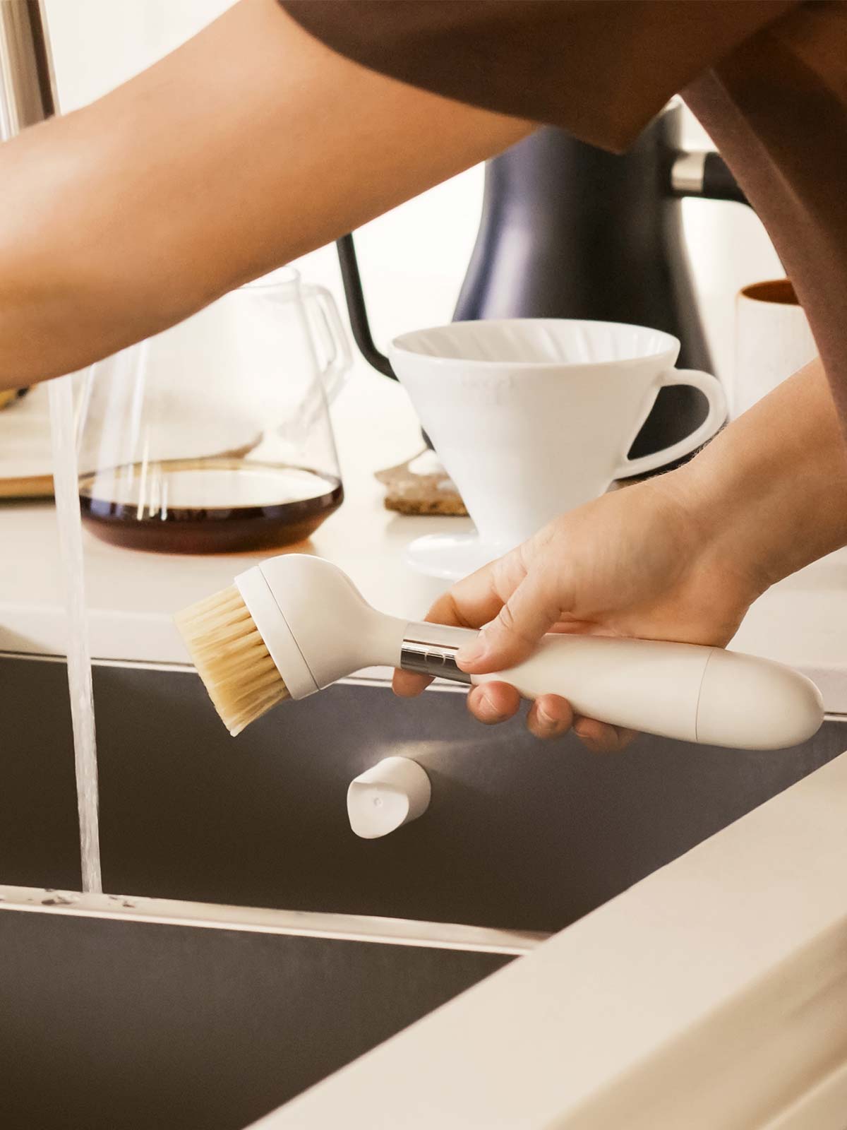 The One Brush V2 Magnetic by Yeseco - Hand holding the white brush over a dark sink, demonstrating the magnetic stand attachment and the sisal fibre brush head.