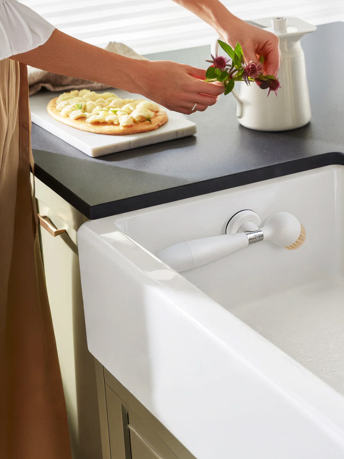 The One Brush V2 Suction by Yeseco - White brush resting on the rim of a white farmhouse sink in a styled kitchen setting, with a person arranging flowers in the background.