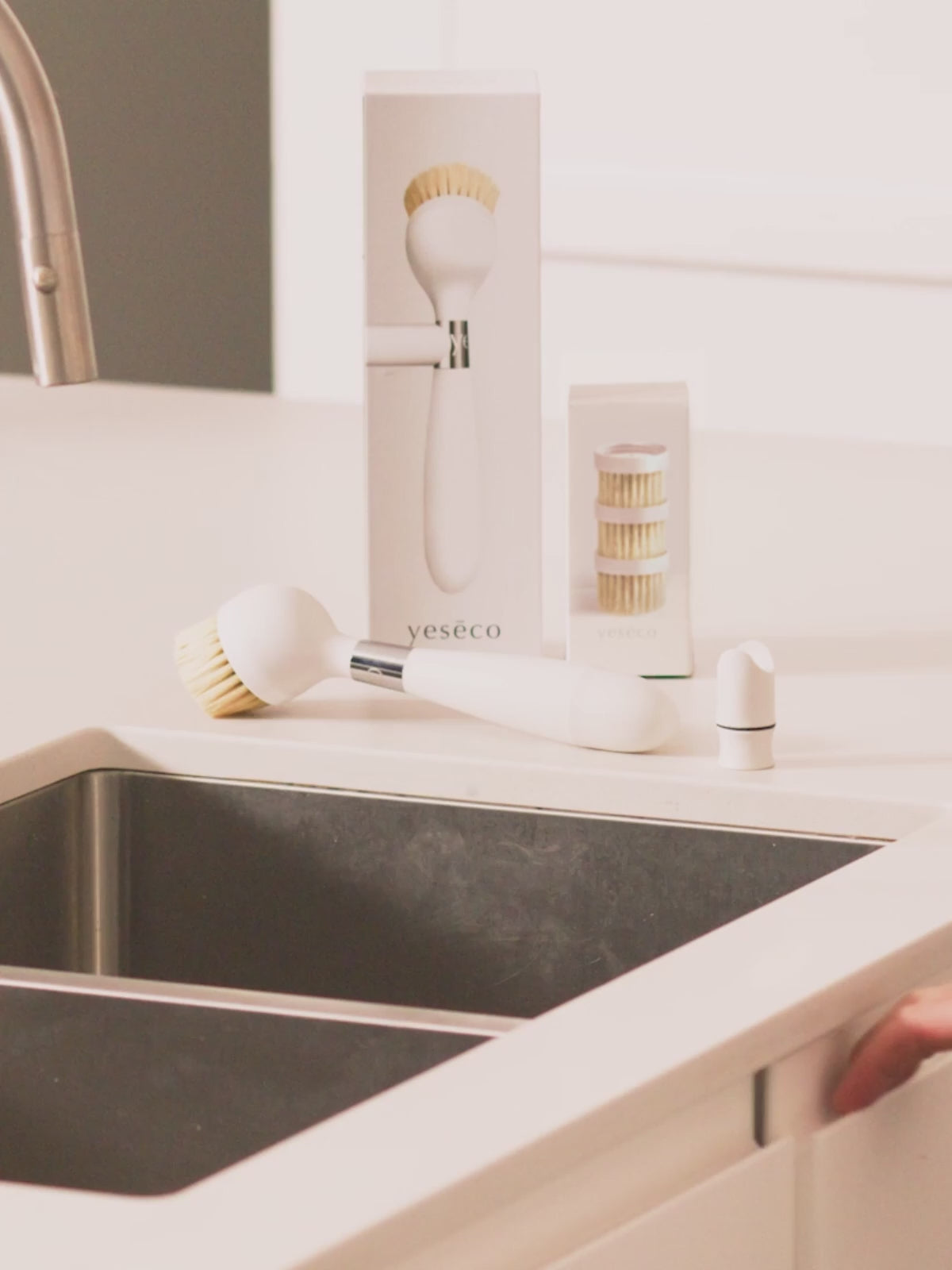 The One Brush V2 Magnetic by Yeseco - White brush resting on the rim of a dark undermount sink in a styled kitchen, showing the magnetic stand clipped to the sink edge.